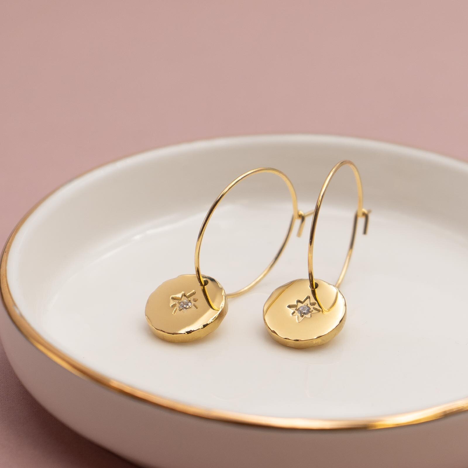 Zwei goldfarbene Creolen mit runden Anhängern aus Edelstahl, Sonnensymbol mit Zirkonia, in einer weißen Schale mit Goldrand
