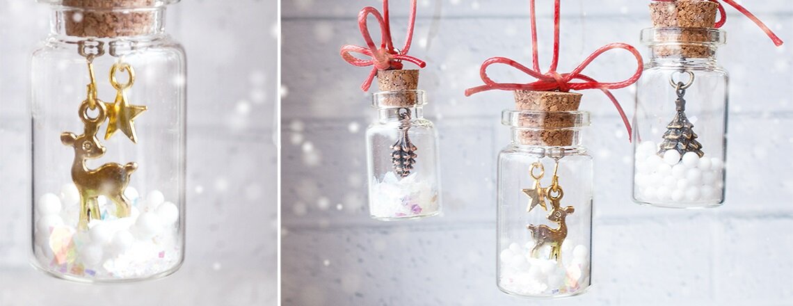 Kleine Glasfläschchen mit Korkverschluss, gefüllt mit weißen Perlen und weihnachtlichen Anhängern wie Rentier, Stern und Tannenbaum, mit roten Schleifen aufgehängt.