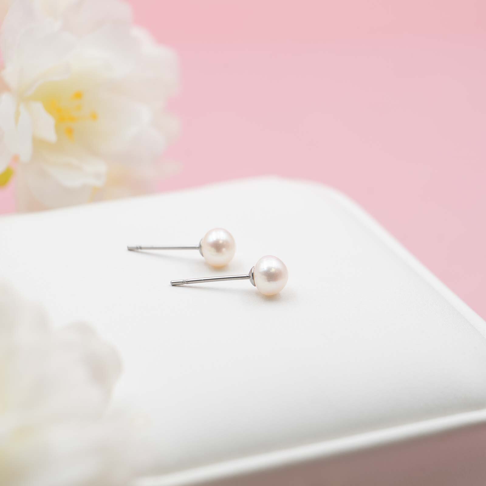 Zwei glänzende, weiße Perlenohrstecker mit silbernem Stift liegen nebeneinander auf einem weißen Schmuckkissen vor rosa Hintergrund und einer weißen Blüte.