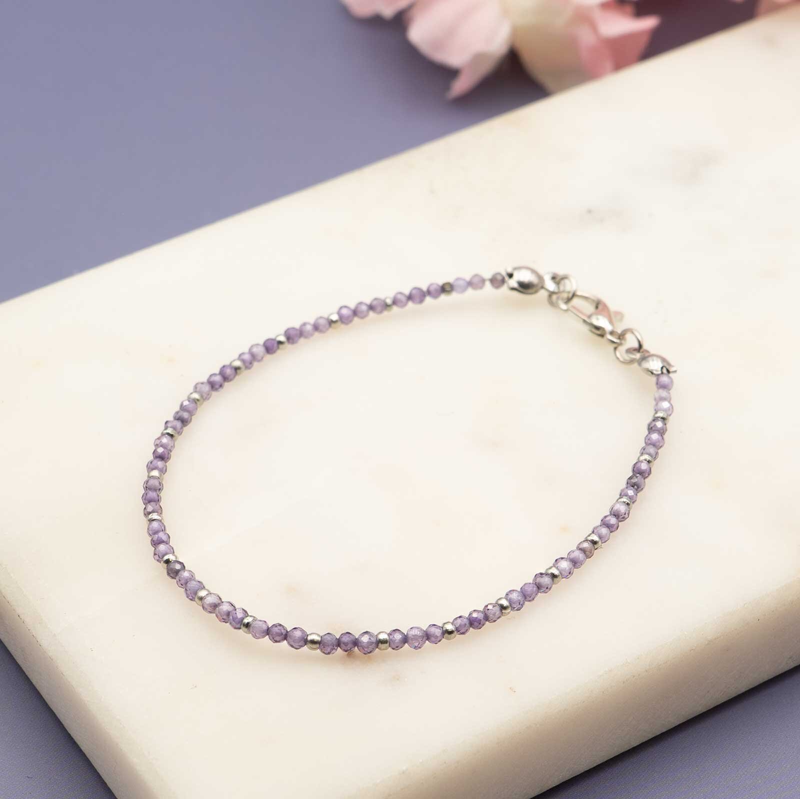 Zartes Armband aus kleinen, facettierten lila Zirkonia- Perlen mit einem silbernen Karabinerverschluss, platziert auf einer hellen Marmorplatte vor violettem Hintergrund.