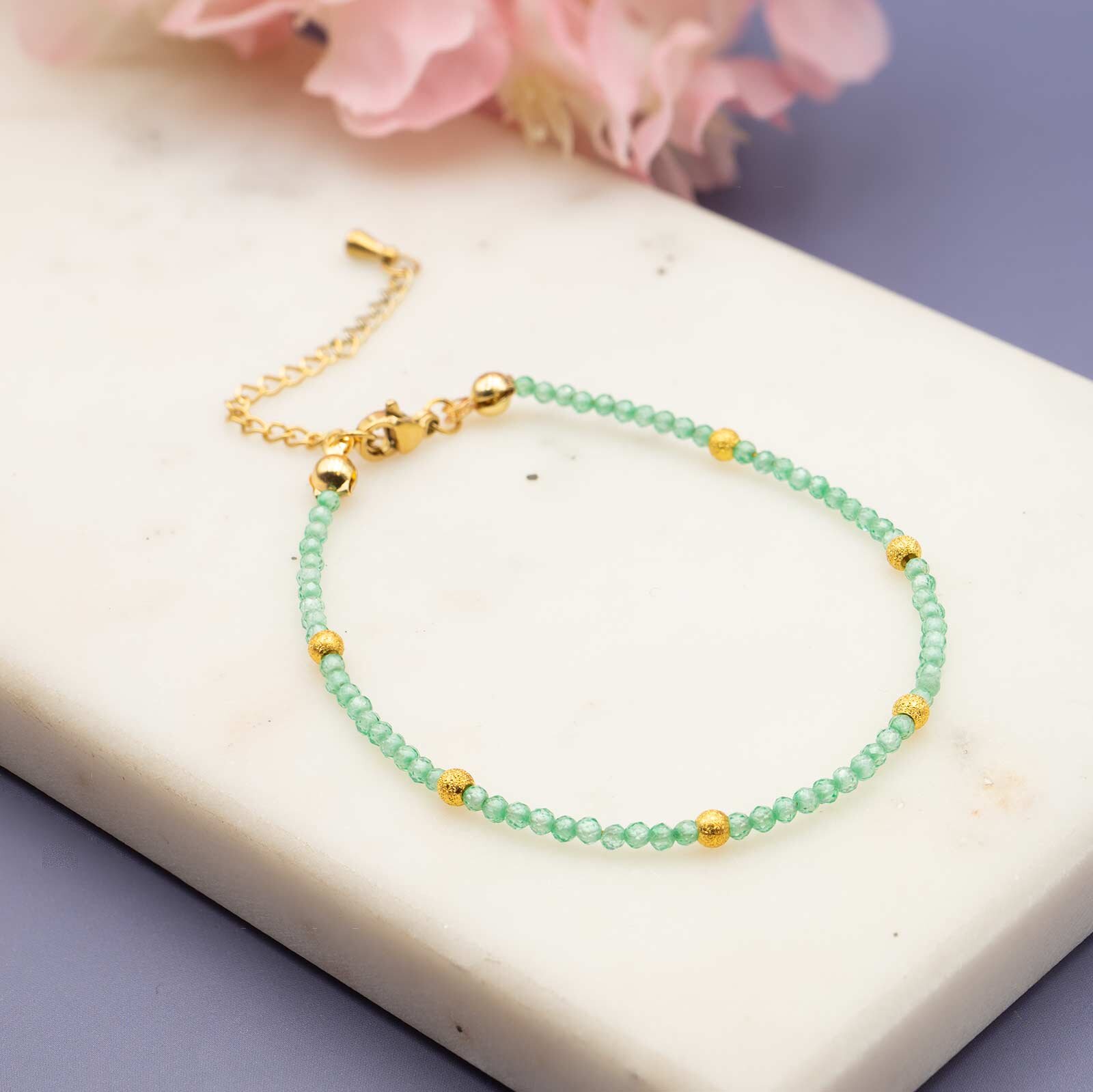 Ein filigranes Armband aus kleinen, grünen Zirkonia- Perlen und fünf gemusterten Goldperlen, das in einem perfekten Kreis auf einer weißen Marmorplatte liegt, mit einem rosa Blumenakzent in der Ecke.