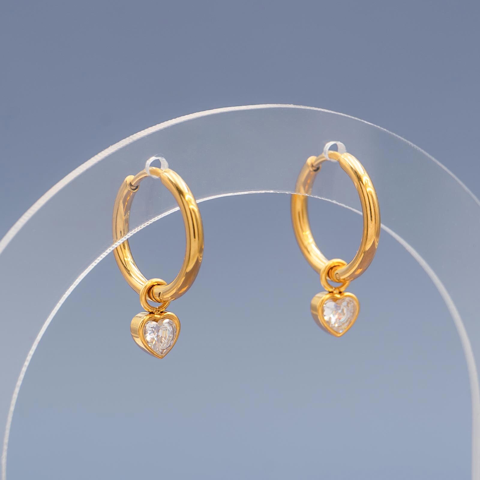 Zwei goldene Creolen-Ohrringe mit funkelnden, herzförmigen Zirkonia-Kristallanhängern hängen an einem transparenten Acryl-Display vor grauem Hintergrund.