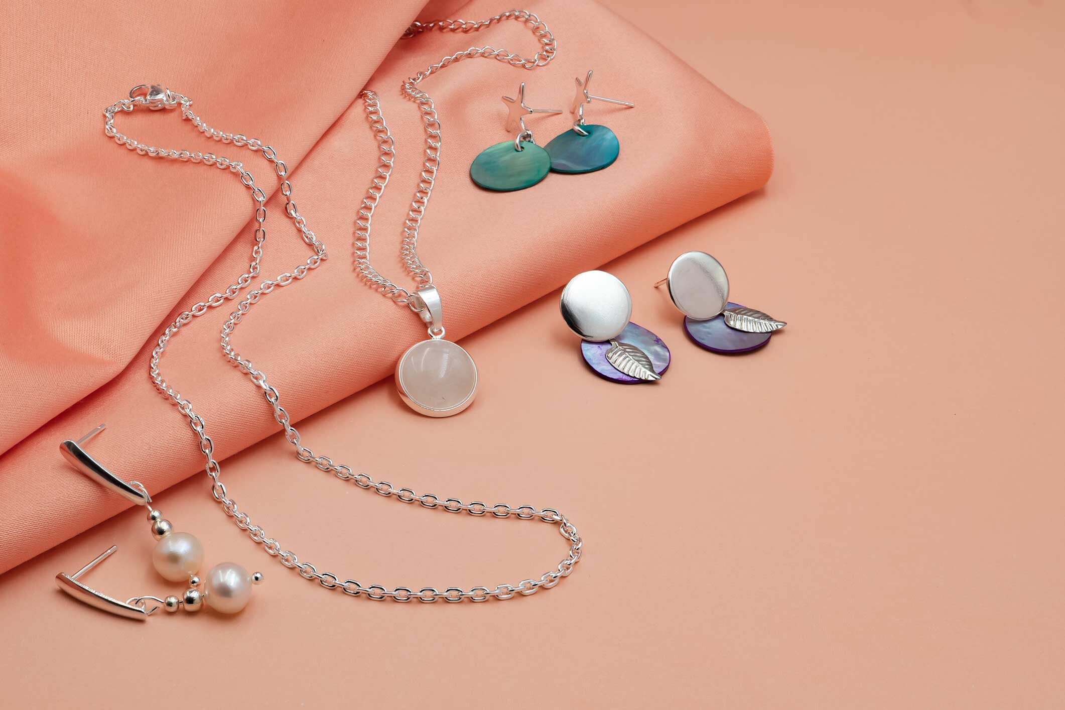 Eleganter Silberschmuck mit Anhängern, Perlen-Ohrringen und Muschel-Elementen auf korallfarbenem Stoff.