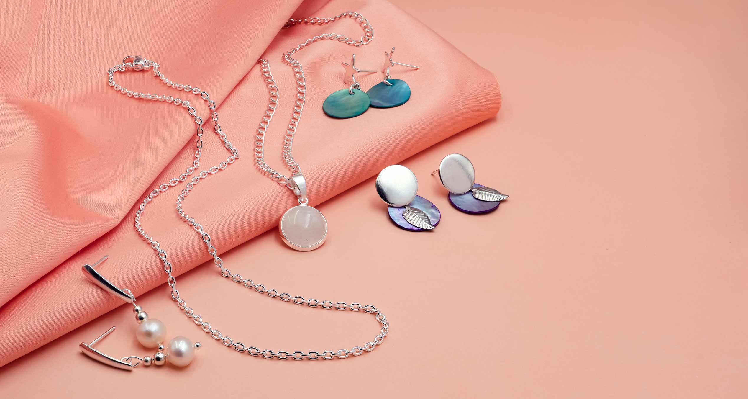 Silberschmuck-Kollektion mit Ketten, Perlen- und Muschel-Ohrringen auf lachsfarbenem Stoff.