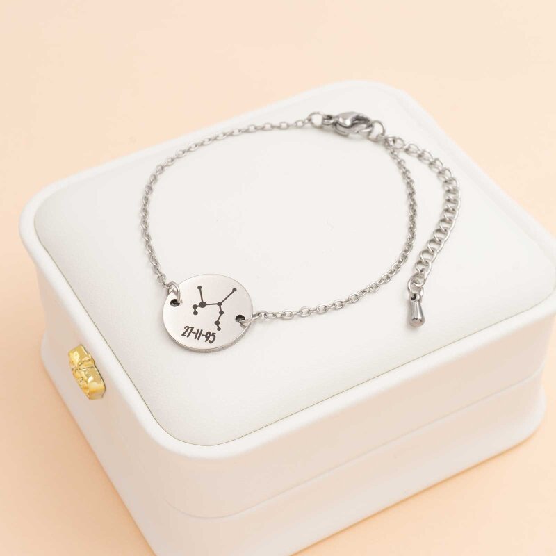 Armband aus feiner platinfarbener Kette mit rundem Edelstahl-Verbinder in der Mitte, graviert mit einem Sternbild und dem Datum 27-11-95, auf einer weißen Schmuckschachtel